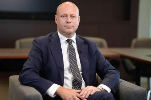 Dan Thomas sitting in a chair looking at the camera wearing a dark suit, white shirt, check tie and a wrist watch.