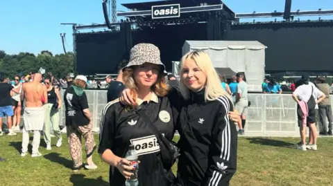 Cerys Hubbard Cerys Hubbard is wearing a bucket hat and black Oasis/ Addidas t-shirt. She is standing with a friend, also wearing a black Addidas top - and behind them we can see they are at one of the recent Oasis gigs.