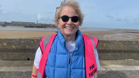 Linzi Foster-Nicholson, volunteer, wears a blue gillet and hi-vis pink jacket which reads 'Poo Patrol'.