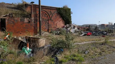 Thomas Ollivier The back of Barrow's Strand station. It is decrepit and has large amounts of ivy growing on the building. The site surrounding is has loads of rubbish dumped outside of it and it has been used as a fly-tipping spot.