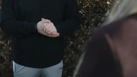 A view of a man from the shoulders down. He is wearing a black top and grey tracksuit bottoms and clasping his hands together as he speaks to a woman with blonde hair and wearing a brown coat, who has her back to the camera.