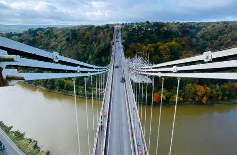 An aerial view of the Clifton Suspension Bridge taken from one of the towers, showing Leigh Woods in the distance and the River Avon below