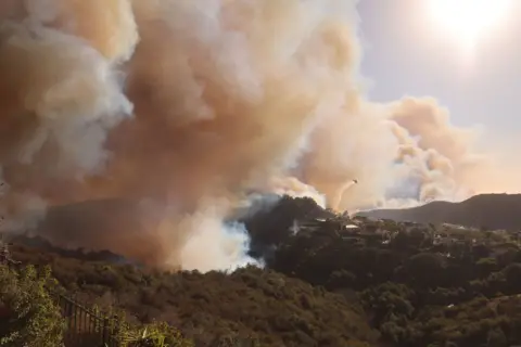 David Swanson/AFP Sebuah helikopter menjatuhkan air ke atas bukit-bukit yang terbakar, dan kepulan asap besar membubung di atasnya, di Pacific Palisades.