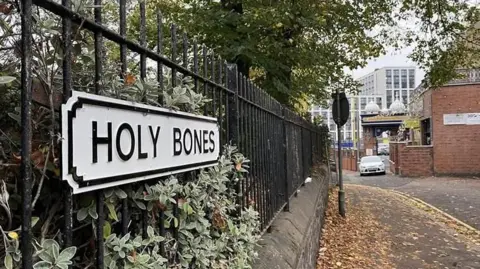 A white and black road sign saying "Holy Bones", attached to a black rail fence. There is a pavement to the right strewn with brown leaves.