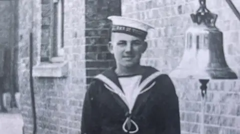 An archive black and white image of Leading Seaman Jack Mantle, dressed in his navy uniform. He is standing next to a bell. He is smiling.