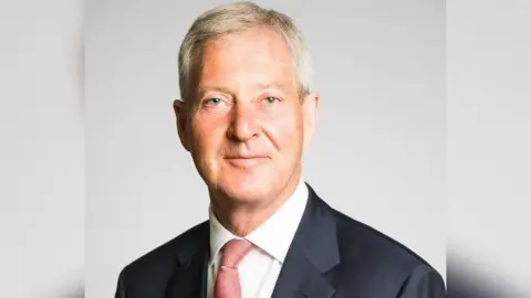 Surrey County Council A headshot of Tim Oliver in which he is looking at the camera. He is wearing a dark suit, white shirt and red tie and is smiling.