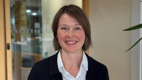 Philippa Read is standing in front of a glass door. She has short brown hair and is wearing a white and black polka dot shirt and a black blazer. 