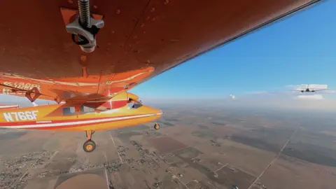 A small orange plane is flying, being towed by another plane in front of it.
