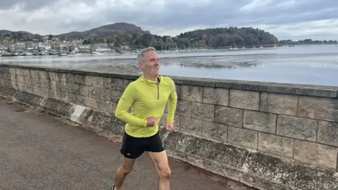Mark Davies in a yellow top and shorts running along The Cob, an embankment in Conwy with a stone wall and the estuary behind - the town of Conwy and mountains in the background.