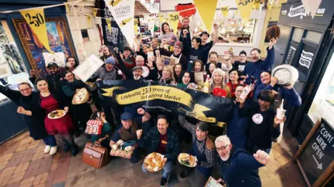 Oxford City Council Traders of the Covered Market gather for a group photo, holding out their wares, such as cooked meals, baked items, milkshakes, flowers, and artwork. Banners and bunting proclaim the 25th-year anniversary.