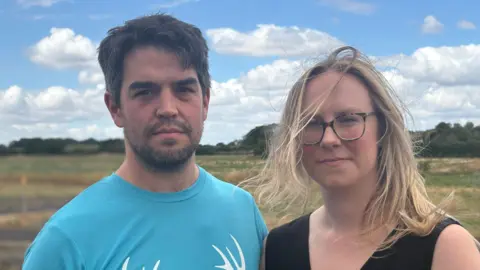 A couple in their 30's standing in front of a field. The man is wearing a light blue t shirt while the woman wears a black top