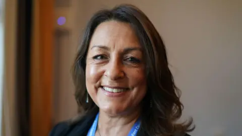 PA Media A woman smiling at the camera. She has long, brown hair and a blue lanyard around her neck.