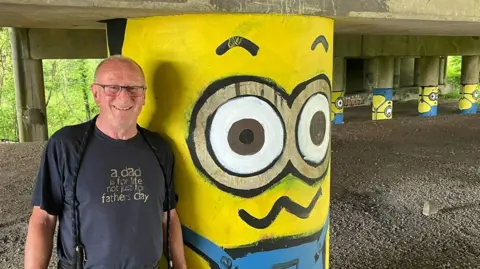 BBC Craig Simpson wearing a navy t-shirt and glasses smiling standing by a motorway stanchions painted to look like a Minion - the yellow cartoon creations featured in the Despicable Me movies.