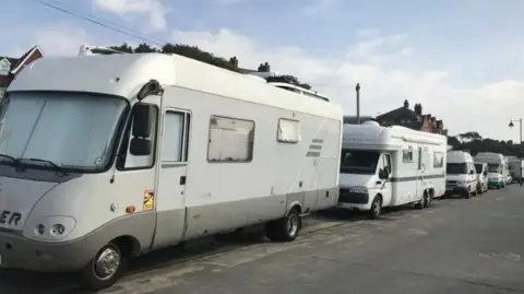 Shannon Eustace/BBC A row of light coloured campervans, parked up bumper to bumper on a road. Above can be glimpsed roofs of buildings. In front is a wide tarmacked road or pavement.