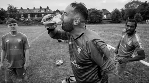 John Bolloten A black and white photo of a man wearing goalkeeper gloves downing a pint of lager at the side of a pitch. Two of his teammates watch on.
