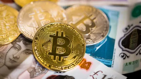 Getty Images A mock-up of what a 'Bitcoin' may look like if it were a physical currency. It depicts a gold coins marked with a B lying scattered on a pile of £10 and £5 notes.