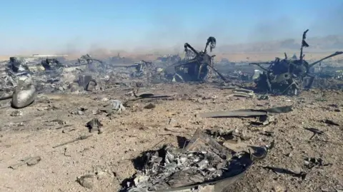 A still image purporting to show the burned wreckage of aircraft destroyed during the US mission to find a stranded airman in Iran. What appears to be two propellers can be seen among scraps of metal and smoke on a sandy ground.