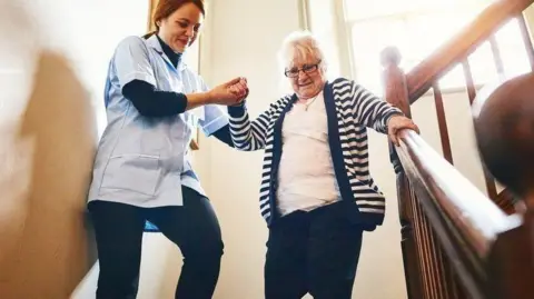 Getty Images Old woman with grey hair is helped down a stair case by a nurse with a window behind them as sunshine pours through