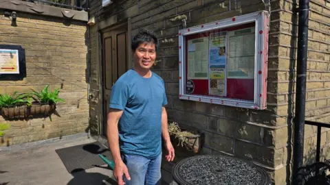 Hayley Coyle/BBC Beng Yau Lim, owner of Poppa Piccolinos, stood outside his restaurant with a menu on display and a wrought iron table.