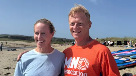 Droneman Helen Glover (L), wearing a light blue top with her blonde hair in a ponytail and smiling. To the right is Joff Machell, wearing a red MND Association top with his arm around Helen and smiling. Behind them is a sandy beach. 