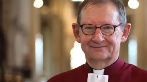 Paul Marriott A smiling Chris Dalliston looks directly at the camera. He is wearing tortoise shell spectacles and has short grey hair. He is wearing a red shirt with a white clerical collar.  He is standing in a cathedral. 
