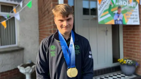 Caolán McConville. Smiling at camera. Wearing navy Special Olympics Ireland jumper with two gold medals hanging down from his neck. 