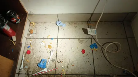 Dirt and rubbish on a floor, including plastic cutlery, drinks cans and food.