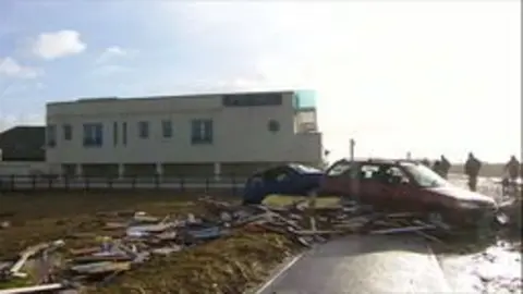 A blue and a red car are abandoned on the grass verge, their windows shattered. They are surrounded by planks of wood, strewn across the pavement and grass. In the background, there is a cream 3-storey building. The sun is shining, reflecting the light on the wet pavement.
