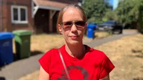 A young woman with blonde hair tied back in a red t-shirt and sunglasses stands outside a house. There is yellowed grass in the background of the image. 