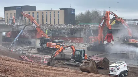 Diggers and trucks around the bridge as it is being demolished. There is what looks like clouds of ash and some rubble in place of the bridge.