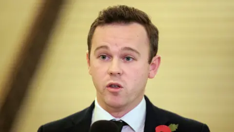 A man with short hair wearing a suit and a poppy standing talking inside. The background is out of focus. 