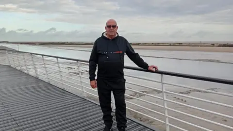 An older man wearing a dark hoodie stands in front of railings overlooking a watery, sandy coastal landscape