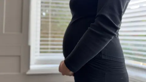 A partial shot of a pregnant woman in profile, dressed in black and standing with her hands joined under her belly. She is in front of a bay window with the outside obscured by Venetian blinds.