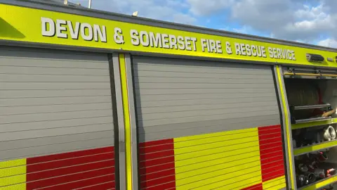 A section of a fire engine with grey roller shutter doors, featuring red and yellow reflective markings. The text 'DEVON & SOMERSET FIRE & RESCUE SERVICE' is prominently displayed. The background shows a partly cloudy sky.