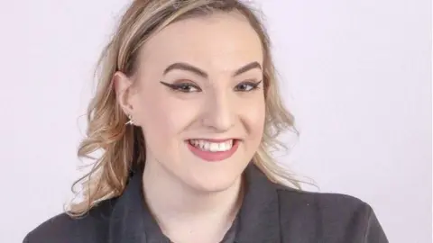 A blonde woman smiles at camera wearing a black top and she has red lipstick on.
