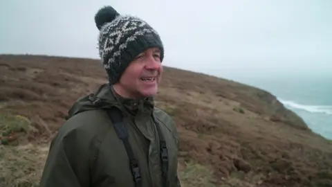 A man stood on a coast path with the sea behind him. It is a misty day and he is wearing a military green coat and a woolly hat, and also has a pair of binoculars around his neck