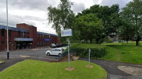 The outside of Pudsey Police Station. A red brick building is on the left of the picture with cars parked opposite. On the right are trees and a grassed area 