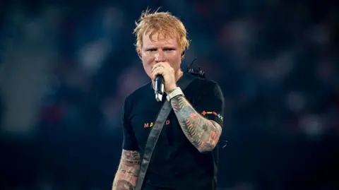 Getty Images Ed Sheeran sings into a microphone. He has a guitar strap over his shoulder and is wearing a black T-shirt with the word "Madrid" across the chest in colourful writing. He has tattoos on his arms and looks quite hot and sweaty.