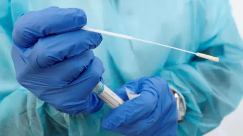 PA Media A nurse clad in sky blue and wearing blue PPE gloves shows a nose swab from a patient in a testing area outside a hospital during the covid pandrmic.