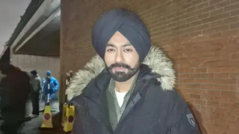Tarsem Singh Jassar wearing a blue turban and blue coat. He has a black beard. He is wearing a white t-shirt, grey shirt and grey top. There is a brick wall underneath a bridge behind him. There are two yellow traffic cones and members of the cast and crew stand in the background.