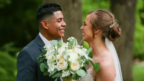 Submitted by Janie Pérez Janie and Alejandro Pérez smile at each other in their wedding attire, Janie in white holding a white rose bouquet