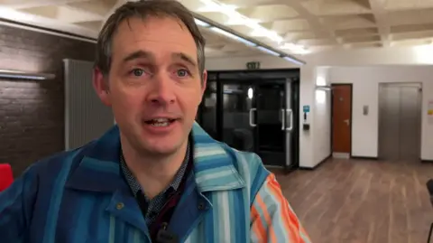 Professor Ed Hawkins is looking at the camera and wearing the climate change stripe jacket which is dark blue, light blue, orange and red.