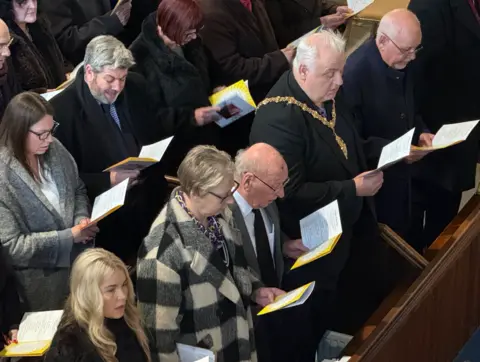 Part of three pews including people holding the order of service.