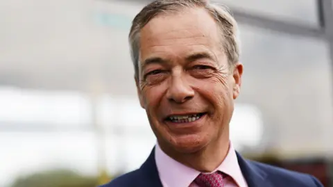 A head and shoulders picture of Nigel Farage, who is smiling and wearing a blue suit and red patterned tie with a pink shirt