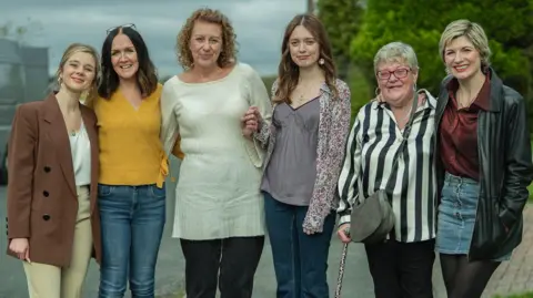 James Stack/Netflix Three female cast members stand smiling with their real-life counterparts. They are on a set that looks like a residential street.