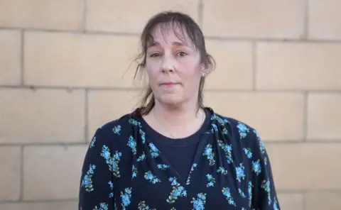 Julie Morgan looking at the camera. She has a fringe and dark hair and is wearing a navy top with blue flowers on, and is standing in front of a brick wall.