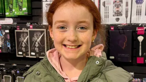 Kelly, nine, with red hair standing in front of darts flights. She is wearing a pink hoodie and green coat. She is smiling. 