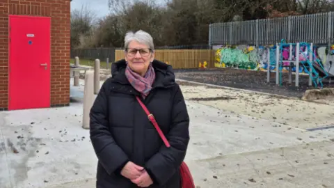 Councillor Janine Howarth stands in front of the renovated facilities on the traveller site, she has a childrens play area to her right and what appeares to be shower blocks on the left