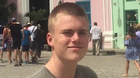 Family photo Christian Hobbs, slightly side on to the camera. He was wearing a khaki coloured t-shirt and is smiling slightly. He had cropped light brown hair. Behind him were cobbles and a mix of different people in summer clothes walking towards a street entry