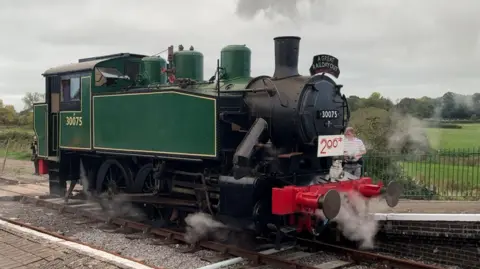 A green and black steam locomotive in steam approaching the platform at Shillingstone